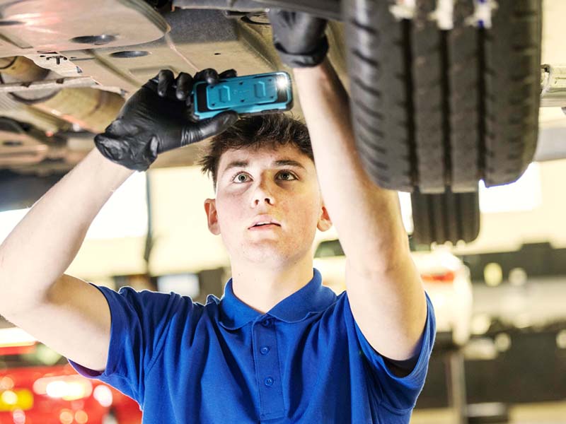 Lookers A Good Deal. Better. Expertise Image Block featuring a male vehicle technician holding a mobile phone under a car during a vehicle service to show a customer.