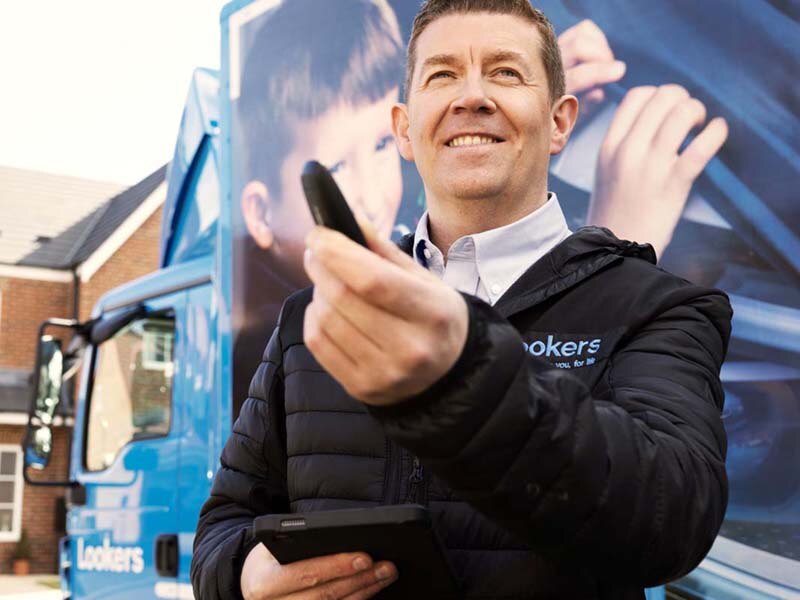 Lookers A Good Deal. Better. Guarantee Image Block featuring a male vehicle delivery driver smiling holding a set of customers car keys being delivered on a residential street.