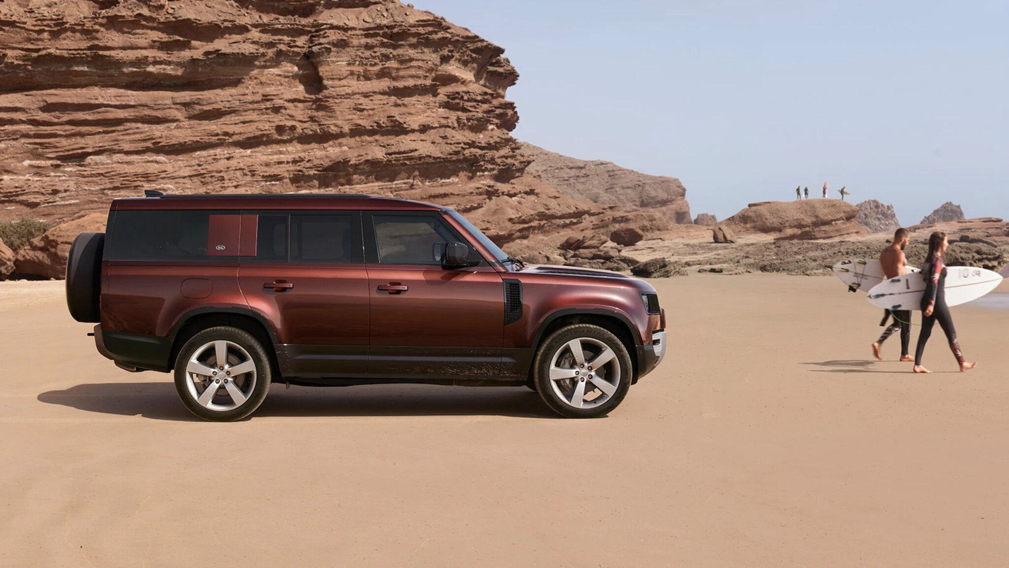 Defender 130 on the beach with 2 surfers walking nearby
